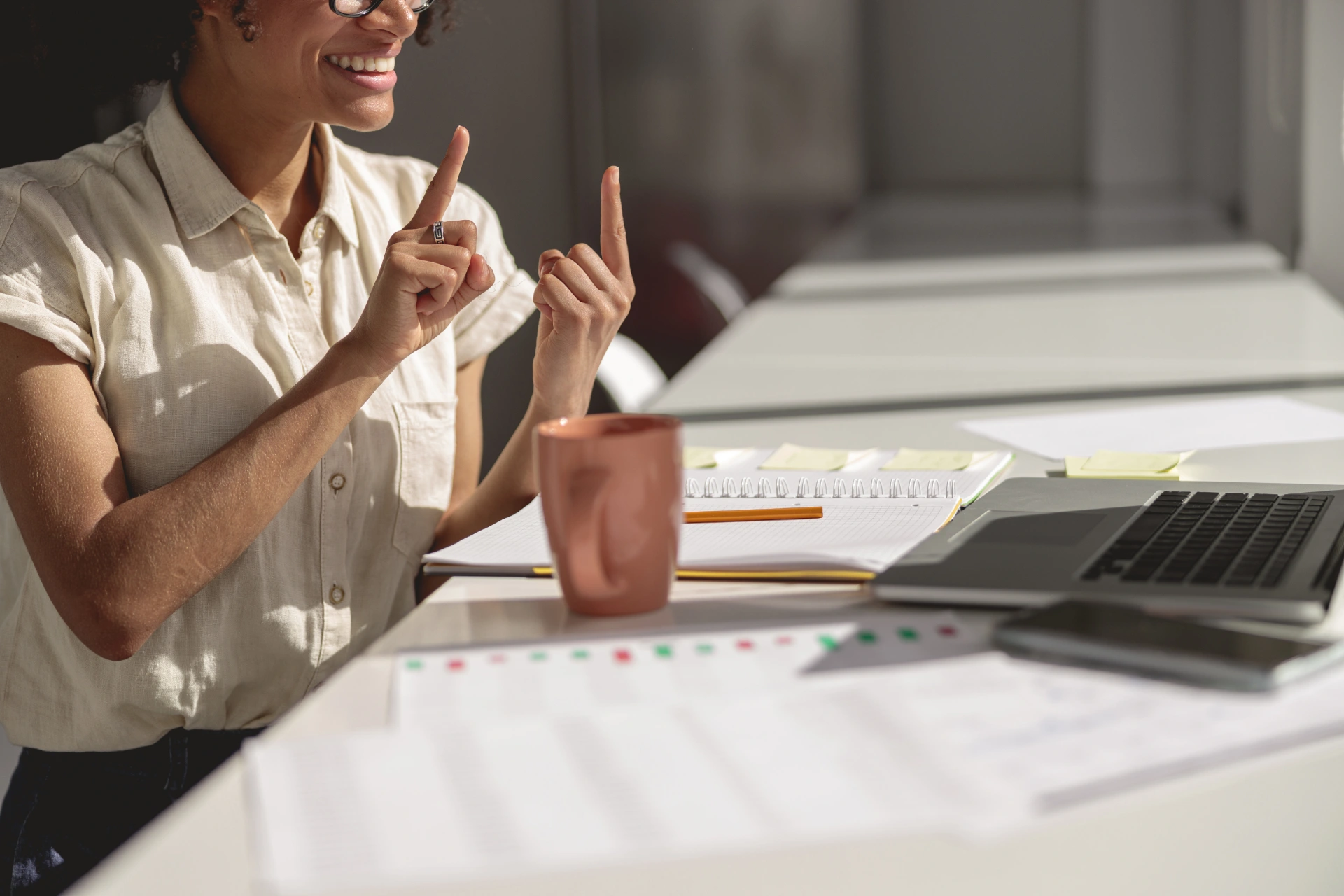 A person sitting at a desk smiles and uses both hands to gesture. On the desk are a laptop, a mug, papers, notebooks, and a pencil.